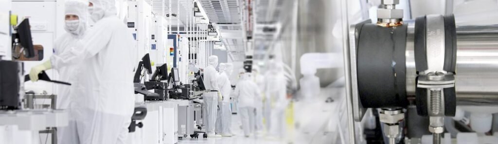 Débitmètre ultrason clamp on installé sur une conduite en acier inoxydable dans une salle blanche industrielle. Au premier plan, un capteur ultrasonore fixé à l’extérieur de la canalisation assure une mesure de débit non intrusif sans contact direct avec le fluide. En arrière plan, plusieurs opérateurs en combinaison intégrale travaillent sur des postes automatisés dans un environnement contrôlé typique de l’industrie pharmaceutique ou des semi conducteurs. Configuration adaptée aux process sensibles nécessitant une mesure de débit ultrason fiable et sans contamination.